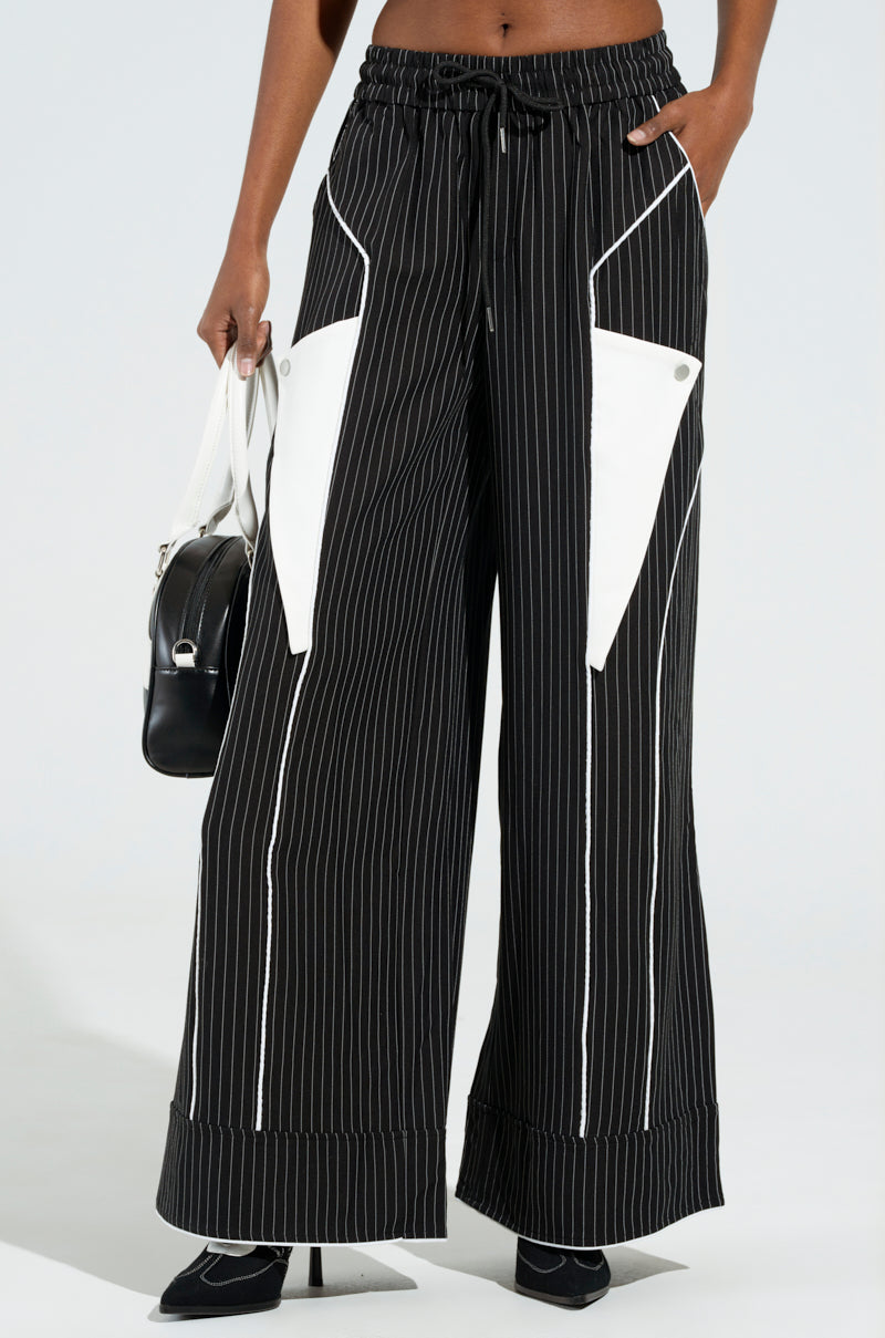 A person wearing the STRICTLY BUSINESS PINSTRIPE PANT, which features a high-rise design, black pinstripes, an elastic waistband with a drawstring, wide-leg silhouette, and white triangular pockets. They are holding a black and white handbag while standing against a plain light background.