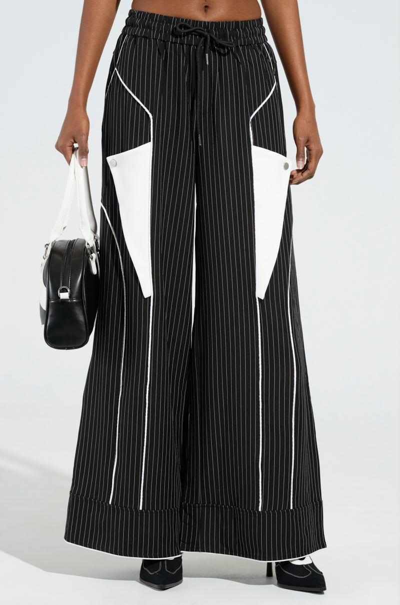 Dressed in the STRICTLY BUSINESS PINSTRIPE PANT, characterized by high-rise black pinstripes and wide legs with prominent white triangular pockets and a drawstring waist, they carry a monochrome handbag and don sleek black shoes against a plain backdrop.