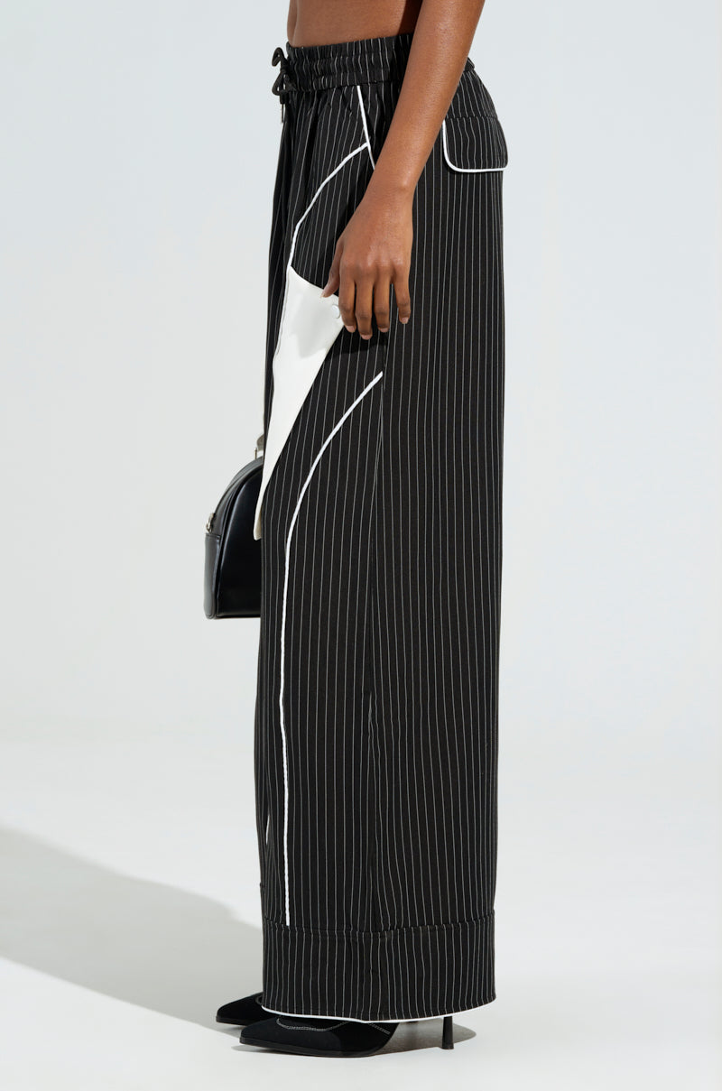 A person is wearing the STRICTLY BUSINESS PINSTRIPE PANT, which features a high-rise design, black pinstripes with white accents, and a relaxed fit with wide legs and a drawstring waist. They complete their outfit with black heeled shoes and hold a sleek black handbag against a plain light-colored background.