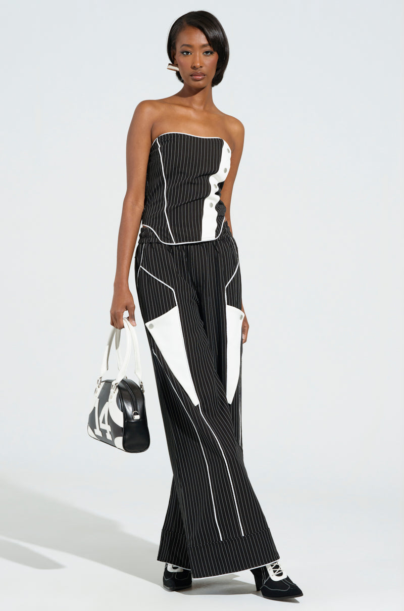 A woman stands on a neutral backdrop wearing the stylish "STRICTLY BUSINESS PINSTRIPE PANT," which features a black and white pinstripe design. Her ensemble includes a strapless top and matching high-rise, wide-leg pants. She completes her look with a black and white bag and black shoes.