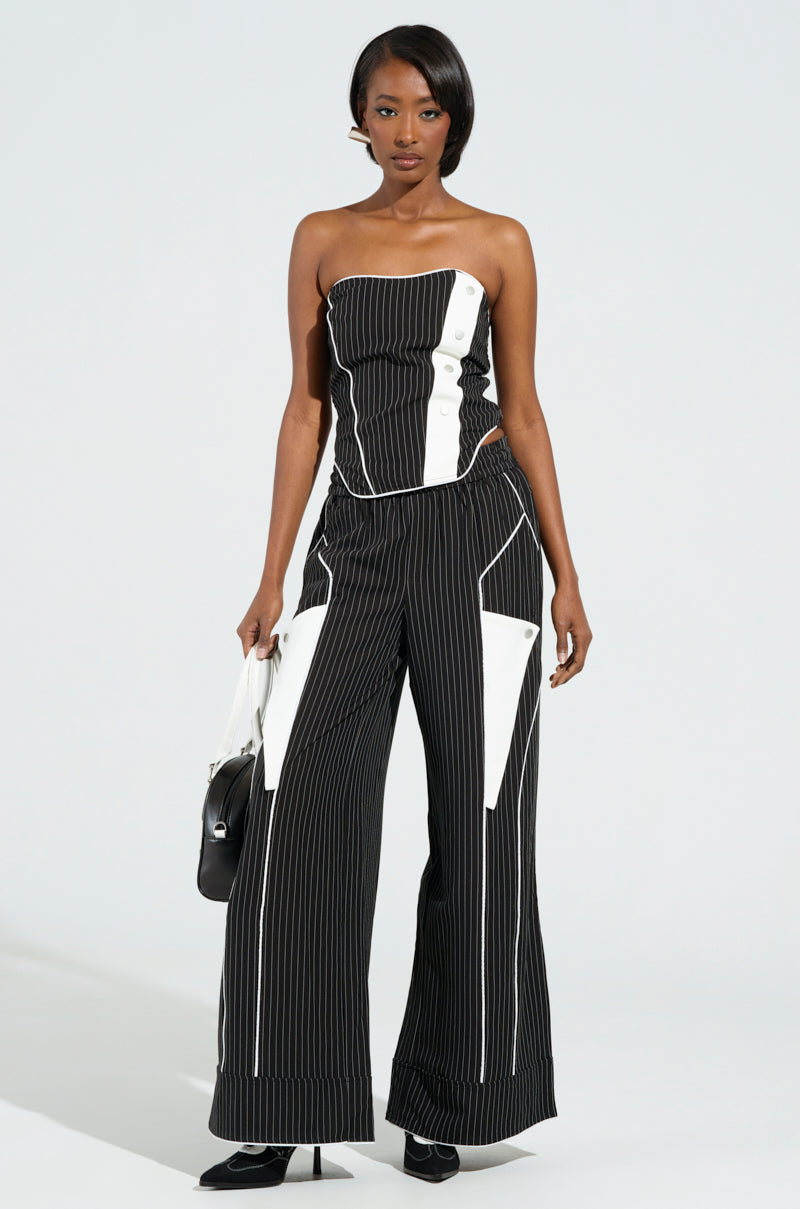 A person wearing the chic Strictly Business Pinstripe Pant, featuring a black and white strapless design with button detailing and high-rise wide-leg style, carries a small black handbag while standing against a plain background.