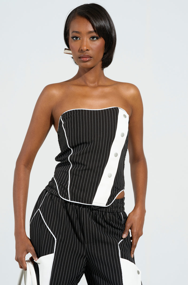 A person models the STRICTLY BUSINESS PINSTRIPE STRAPLESS TOP, a corset-inspired, strapless black and white pinstripe piece featuring button details, paired with matching pants against a plain background, highlighting the elegance of its stretchy fabrication and complemented with hoop earrings.