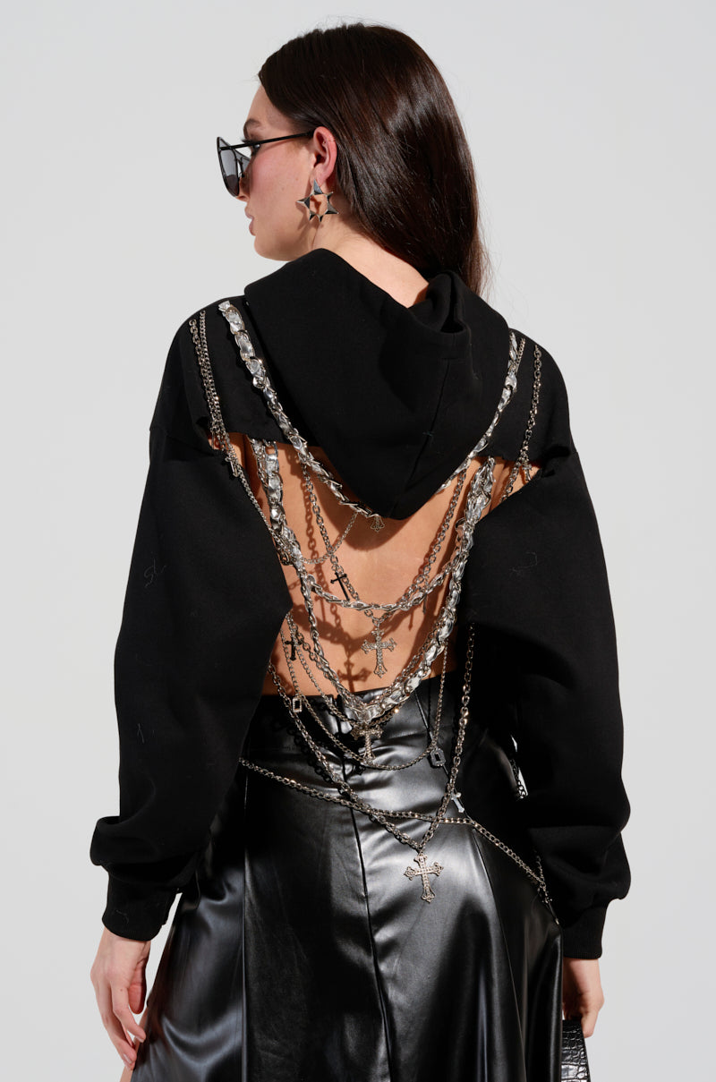 A person stands with their back to the viewer, dressed in the CHAINS FOR YOU HOODIE which features an open back embellished with faux silver metallic chains. They pair it with a shiny black skirt and dark sunglasses, creating an edgy look accentuated by dangling cross pendants.