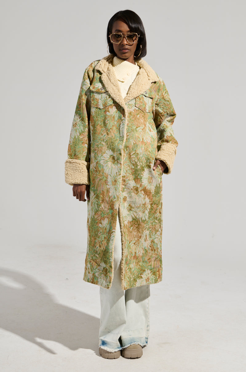 Against a plain background, a person exudes a stylish and confident vibe wearing the HERE COMES TROUBLE SHERPA LINED TRENCH. The long coat features a floral pattern with beige sherpa-lined cuffs and collar. Paired with sunglasses, a white shirt, and light-colored pants, they have their hands in pockets.