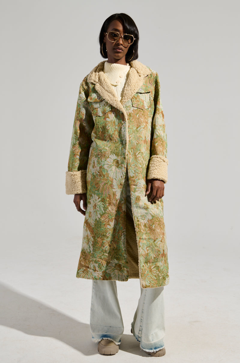 A person wearing sunglasses poses in a "Here Comes Trouble Sherpa Lined Trench," which features a long, brocade-patterned design with plush collars and cuffs. The trench coat boasts a maxi longline silhouette worn over a cream sweater and light blue jeans, completed with beige shoes. The background is plain and light-colored.