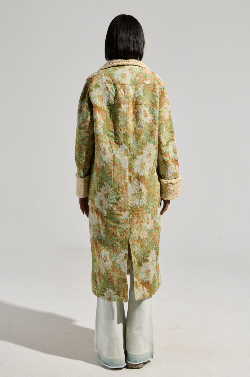 A person is seen from behind sporting the HERE COMES TROUBLE SHERPA LINED TRENCH, which boasts a floral brocade design in light green and beige hues. This maxi longline silhouette coat is complemented by wide cuffs and matched with light-colored, loose-fitting pants, all set against a neutral and minimalist backdrop.