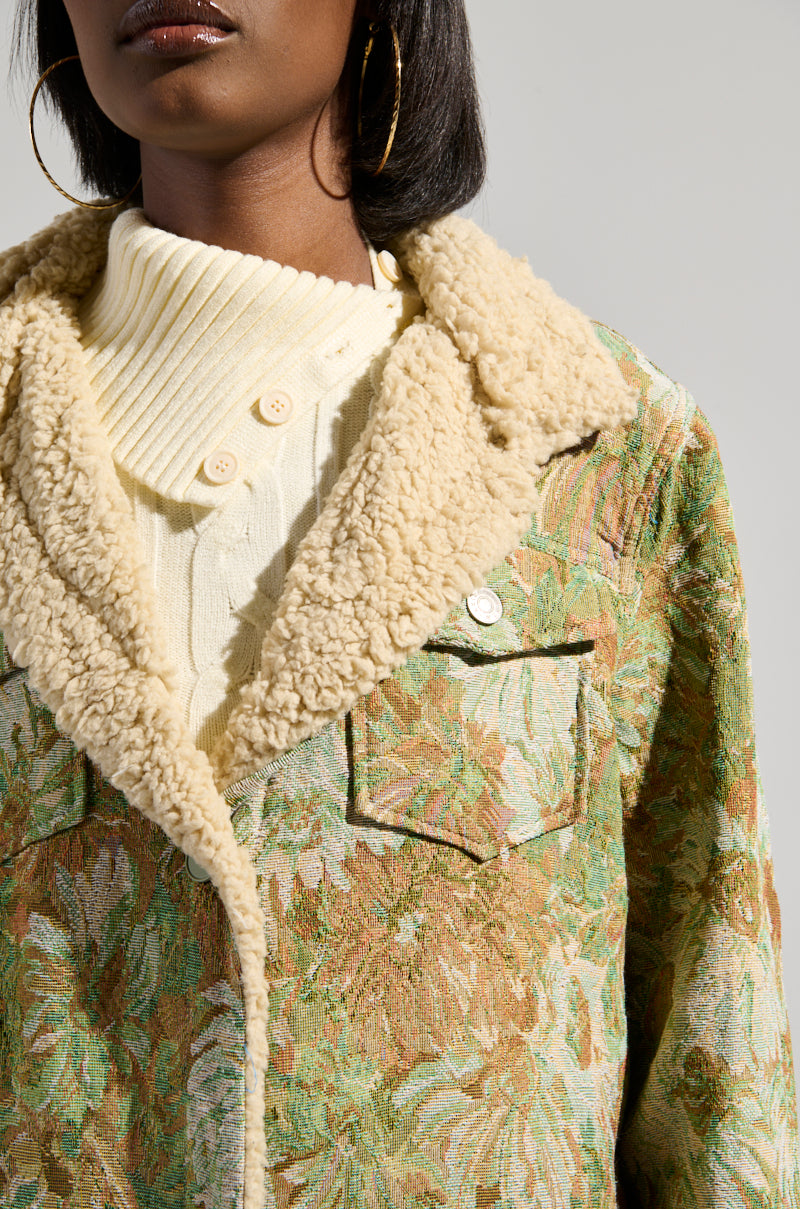 A person is wearing the "Here Comes Trouble" sherpa-lined trench, featuring a plush collar and floral pattern, over a cream ribbed sweater with buttons. They have gold hoop earrings and dark shoulder-length hair. The background is a plain, light color.