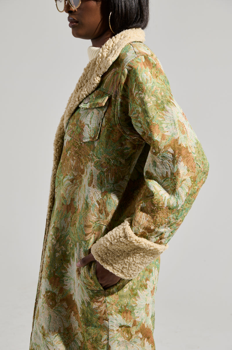 A person wearing the HERE COMES TROUBLE SHERPA LINED TRENCH, adorned with a brocade pattern, steps into the scene. This long coat showcases a floral design and is lined with plush, cream-colored sherpa on the interior and cuffs. A convenient chest pocket adds functionality. The ensemble is finished with glasses perched on their nose and a white turtleneck, all set against a neutral backdrop.