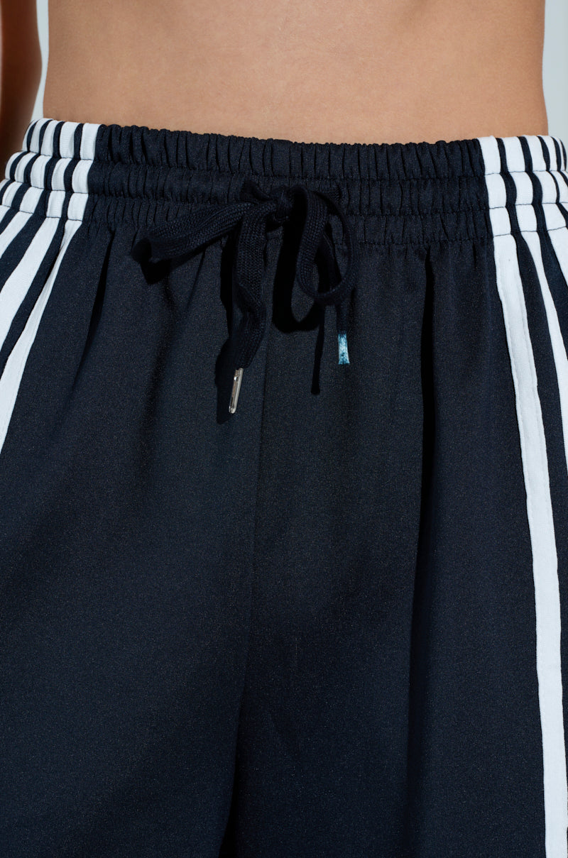A close-up of the JUMP AROUND WIDE LEG PANT in black, featuring striking white stripes down the sides. An elastic waistband with a central drawstring provides comfort, while a neutral background accentuates their sleek style.