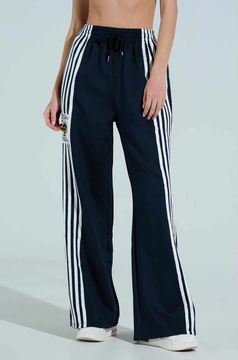 Against a plain background, someone stands in sporty chic black JUMP AROUND WIDE LEG PANTS. These high-waisted track pants, adorned with white vertical stripes and a leg patch, pair perfectly with white sneakers for a trending high-rise look.