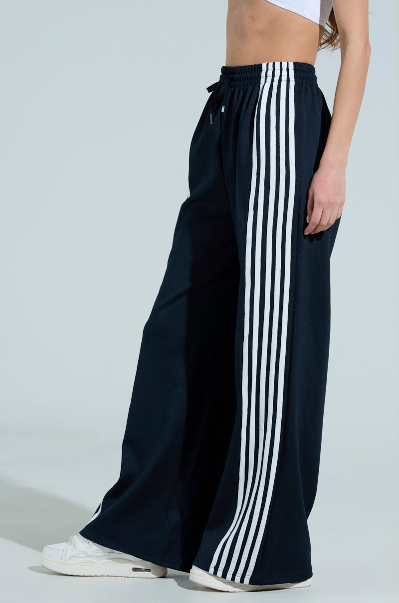 A person exudes sporty chic against a light gray background, wearing JUMP AROUND WIDE LEG PANTs featuring vertical white stripes. The high-rise design pairs well with a white crop top, and crisp white sneakers complete their look. Only part of the upper body is visible.