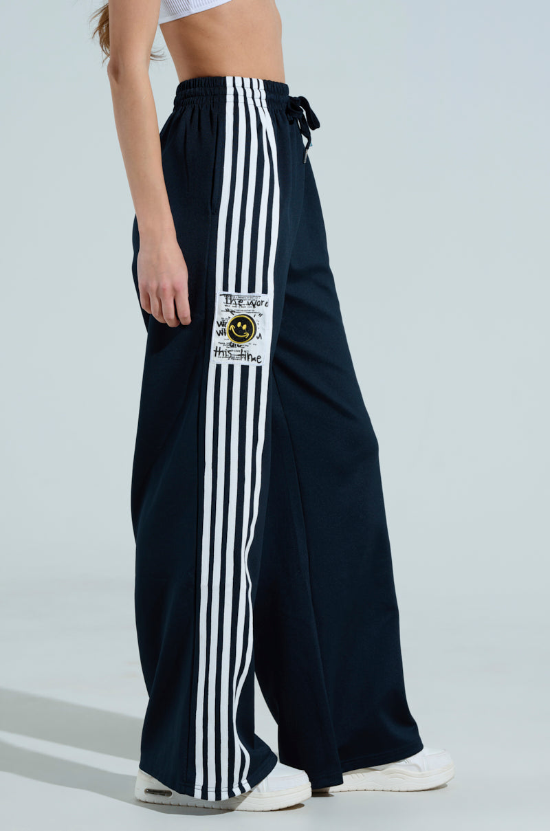 A person showcases the JUMP AROUND WIDE LEG PANT in dark blue, featuring white side stripes and a smiling face patch with "Be your own sunshine" text, embodying sporty chic. These high-rise pants are elegantly paired with white sneakers.
