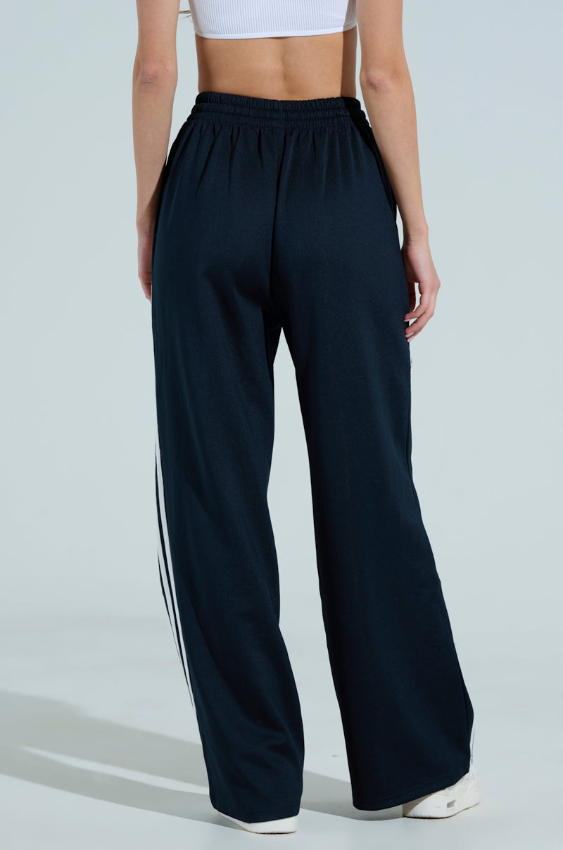 A person embodies sporty chic in the JUMP AROUND WIDE LEG PANT, featuring wide-leg black track pants with white stripes and a white cropped tank top, confidently standing on a light-colored surface, facing away from the camera.