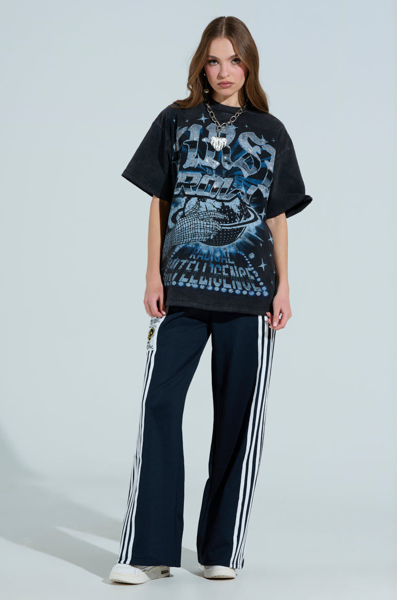 A woman with long hair wears a black graphic t-shirt and JUMP AROUND WIDE LEG PANT, both featuring a smiley face logo. She stands against a light background, completing her look with white sneakers and layered necklaces.