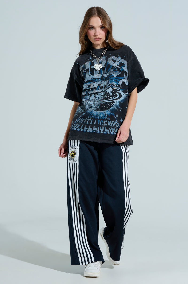 A woman showcases a black graphic tee with bold blue and white text, paired with JUMP AROUND WIDE LEG PANT featuring bold white vertical stripes, a leg patch, and a shirred elastic waistband. She accessorizes with layered necklaces and completes her look with white sneakers.