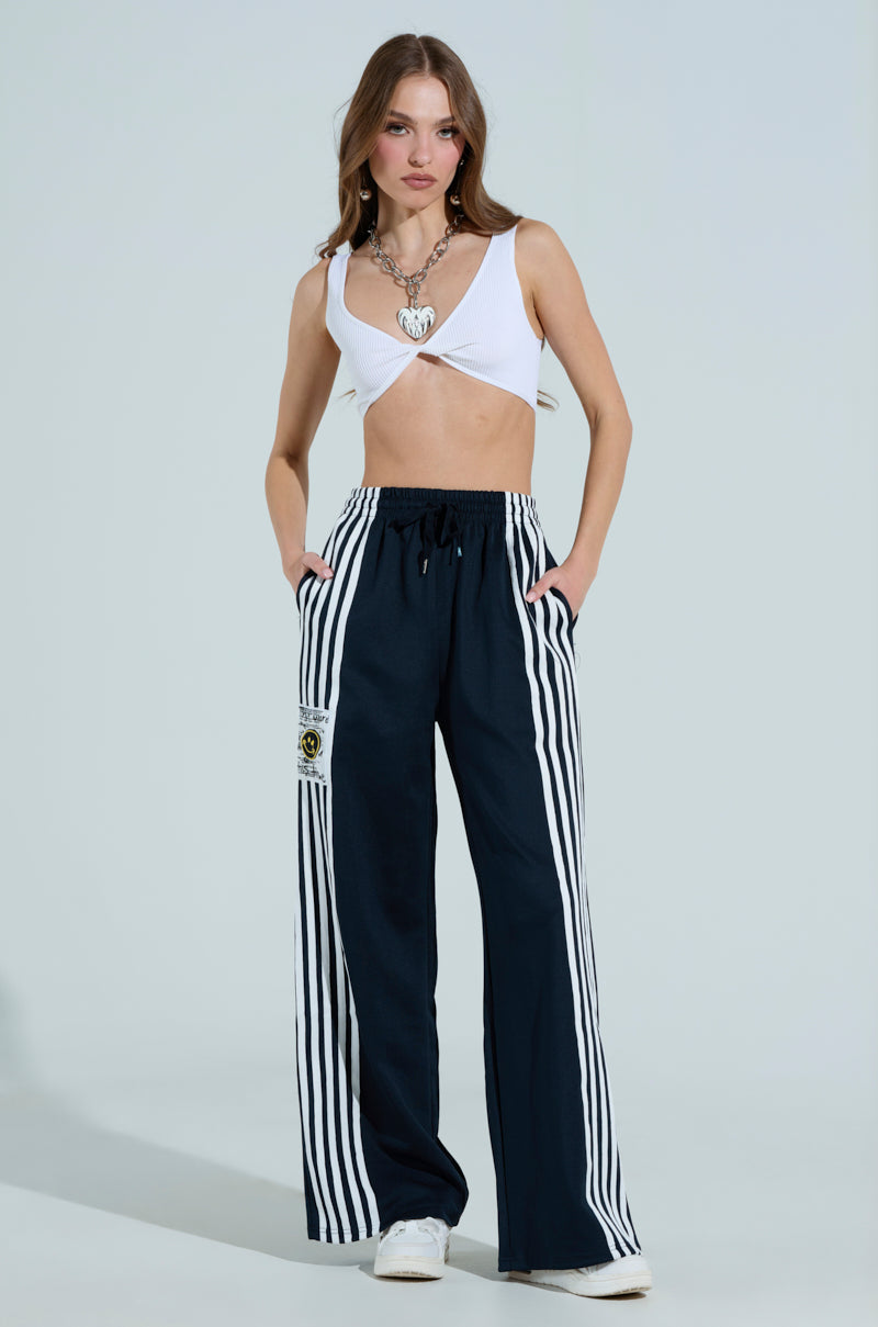 A woman sports a chic look in a white crop top with unique cut-outs, paired with the JUMP AROUND WIDE LEG PANT featuring white side stripes. She stands with hands in pockets, layered necklaces, and crisp white sneakers complete her style.