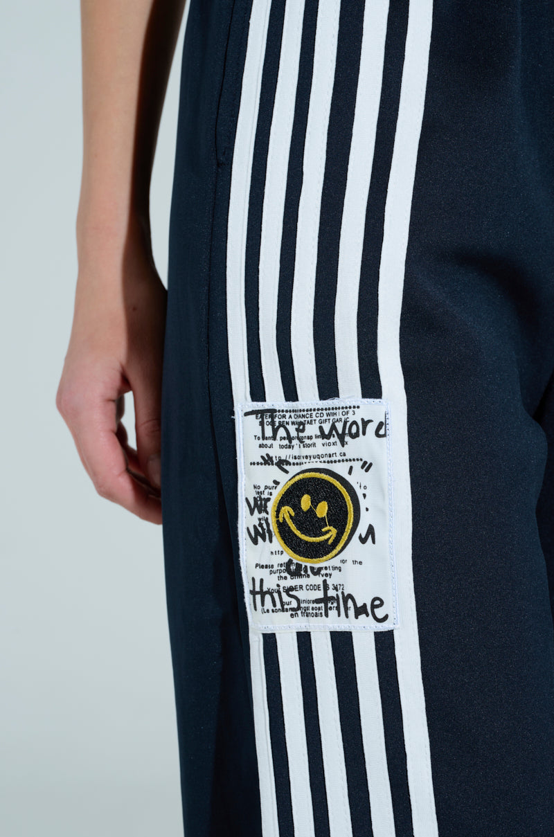 Close-up of someone wearing JUMP AROUND WIDE LEG PANT in navy blue with white side stripes, an elastic drawstring waistband, and a high-rise fit. A fabric patch displays a black smiley face and handwritten text, including "the word" and "this time.