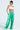 Wearing a white off-shoulder crop top, JUMP AROUND WIDE LEG JOGGER IN GREEN featuring white stripes, and yellow-tinted sunglasses, their tattooed chest and arms exude confidence against a plain backdrop, capturing an effortless athleisure vibe.