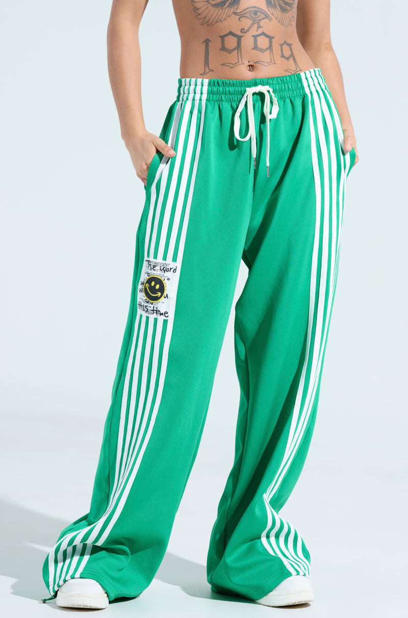 The individual wears JUMP AROUND WIDE LEG JOGGER IN GREEN with white stripes and a yellow smiley patch, showing off a tattooed torso above the waistband. Crisp white sneakers complement the look against a plain light blue backdrop.