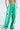 The individual wears JUMP AROUND WIDE LEG JOGGER IN GREEN with white stripes and a yellow smiley patch, showing off a tattooed torso above the waistband. Crisp white sneakers complement the look against a plain light blue backdrop.