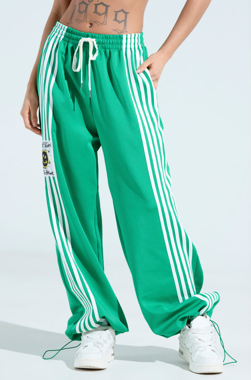 A person is sporting the JUMP AROUND WIDE LEG JOGGER IN GREEN, featuring white vertical stripes, a drawstring waist, and a smiley face patch. They pair it elegantly with white sneakers and flaunt a tattoo above the waistband, perfectly embodying an athleisure vibe.