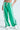 A person is sporting the JUMP AROUND WIDE LEG JOGGER IN GREEN, featuring white vertical stripes, a drawstring waist, and a smiley face patch. They pair it elegantly with white sneakers and flaunt a tattoo above the waistband, perfectly embodying an athleisure vibe.