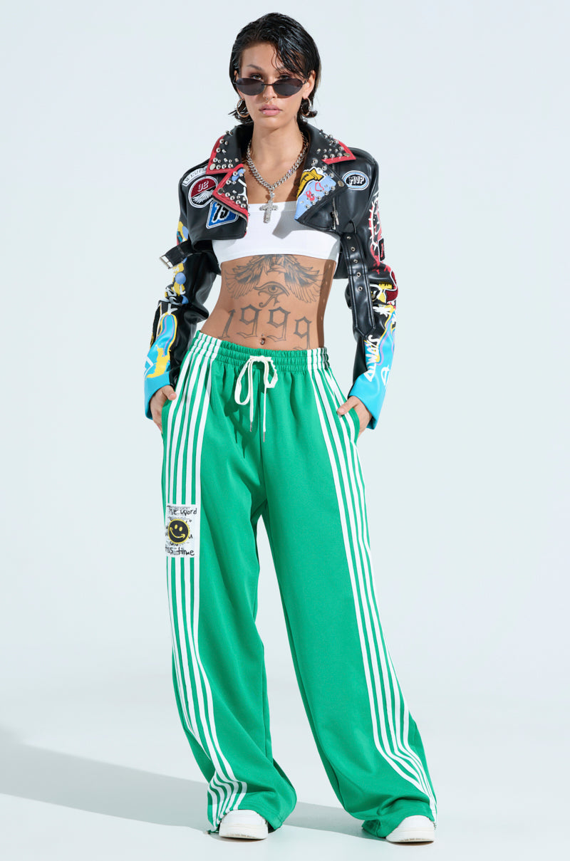 Wearing a black and multicolored cropped leather jacket, white crop top, and the JUMP AROUND WIDE LEG JOGGER IN GREEN with white stripes, they exude athleisure chic. Donning sunglasses and short dark hair, they confidently pose against a plain background.