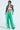 Wearing a black and multicolored cropped leather jacket, white crop top, and the JUMP AROUND WIDE LEG JOGGER IN GREEN with white stripes, they exude athleisure chic. Donning sunglasses and short dark hair, they confidently pose against a plain background.