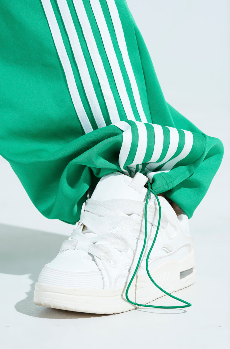 Close-up of a white sneaker with green accents paired with the JUMP AROUND WIDE LEG JOGGER IN GREEN. The right pant leg is tucked into the shoe, showcasing the athleisure style and contrasting colors.