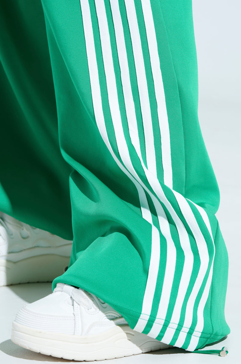 A person sports the JUMP AROUND WIDE LEG JOGGER IN GREEN with white stripes and white sneakers, exuding an athleisure vibe as the loose fit of the joggers partially covers the sneakers against a light, plain background.