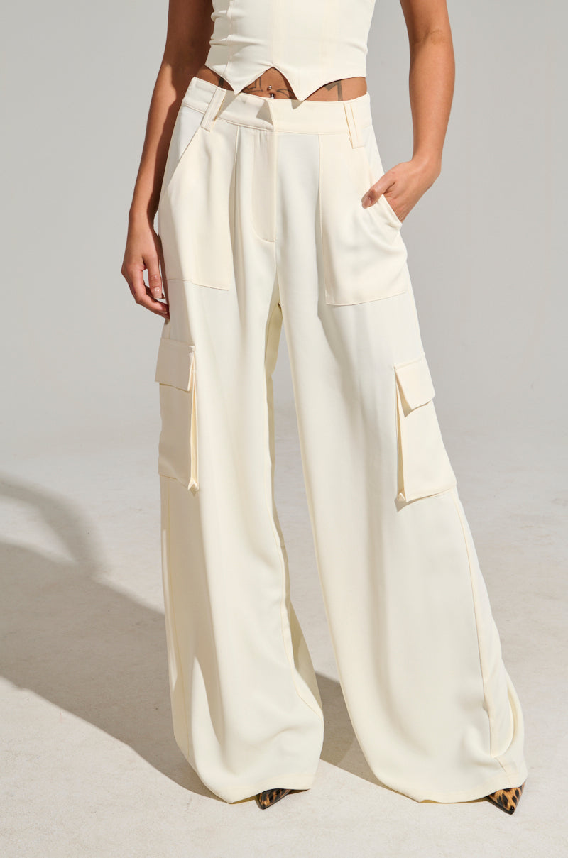 A person is wearing the DRIFT AWAY TROUSER, a minimalist piece featuring high-rise wide-leg cream cargo pants with practical cargo-style pockets, paired with a matching sleeveless crop top. Standing against a plain light background, they have one hand casually tucked into a pocket.