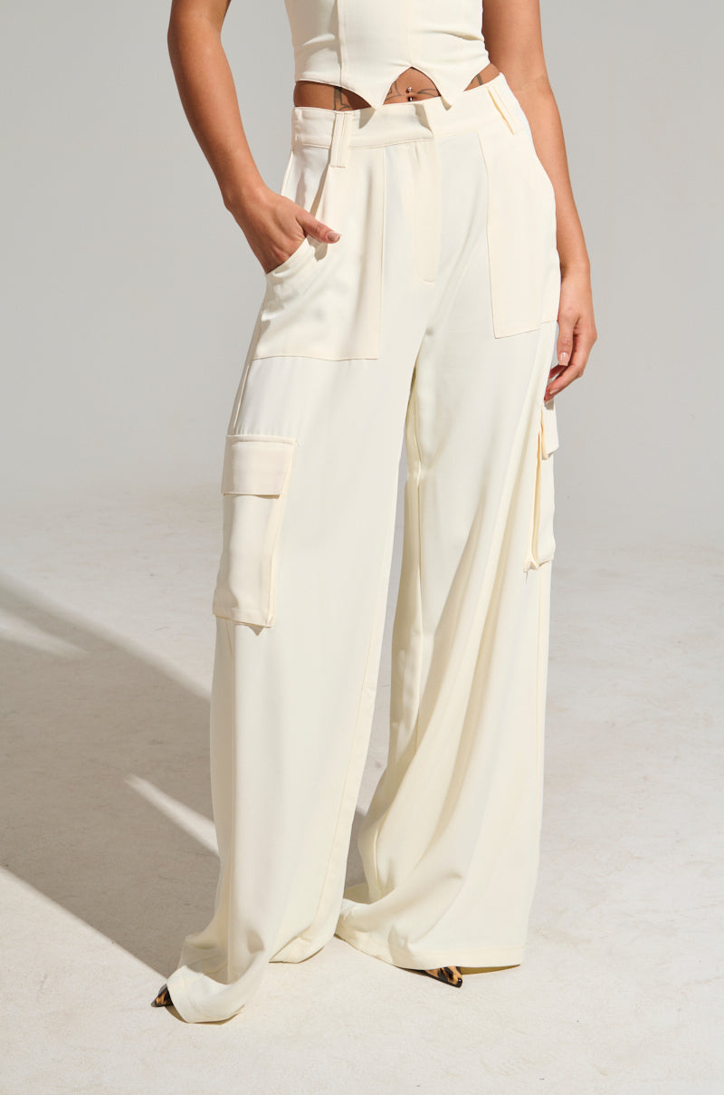 A person dons a minimalist ensemble featuring the DRIFT AWAY TROUSER, cream-colored high-rise wide-leg trousers with cargo-style pockets. The matching cropped top complements the modern, relaxed fit. One hand rests casually in their pocket against a neutral, soft gray backdrop.