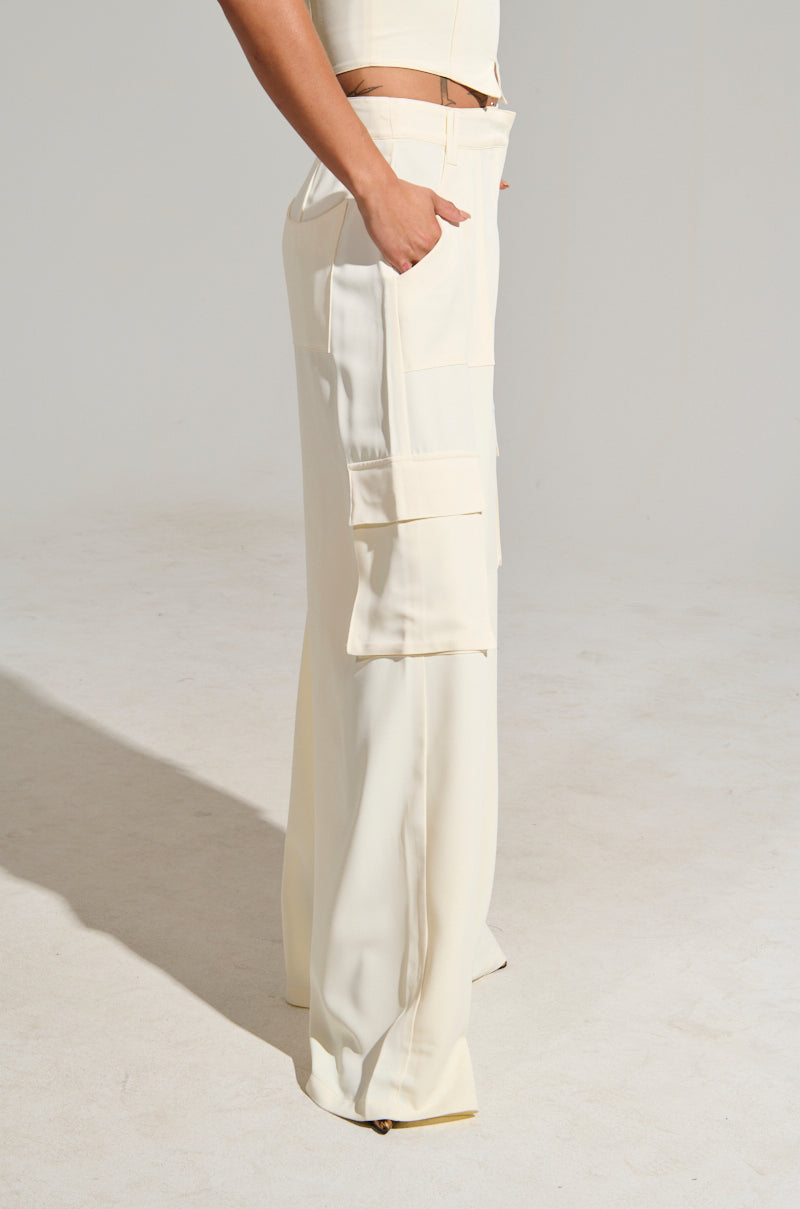 A person is dressed in a minimalist ensemble featuring the cream-colored DRIFT AWAY TROUSER, which boasts a high-rise, wide-leg design with cargo-style pockets. In a neutral environment, the scene is well-lit to beautifully highlight the fabric's texture as they casually place one hand in a pocket.