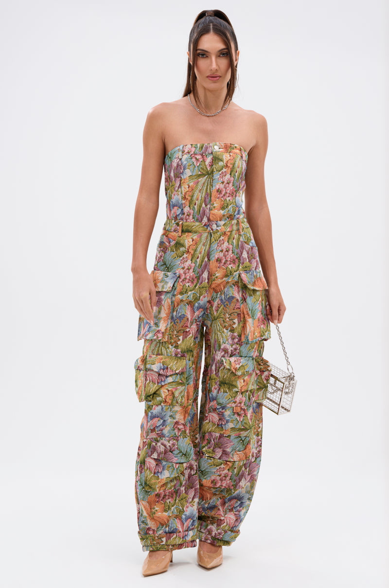 A woman in the STAR OF THE SHOW TAPESTRY JUMPSUIT, featuring a strapless design, wide legs, and a vibrant leaf pattern, stands against a white background holding a boxy handbag and wearing nude heels.