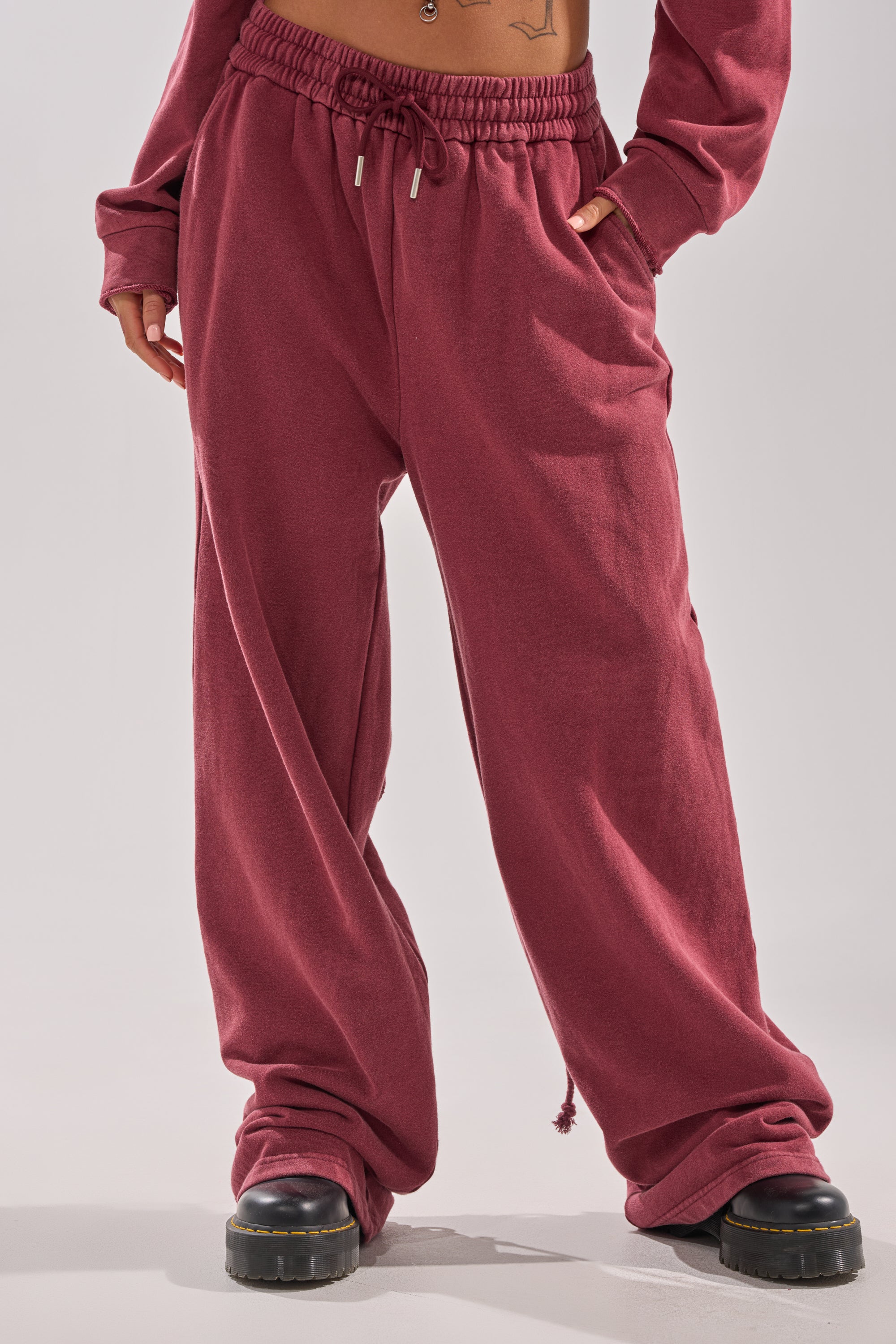 A person wearing the LACED UP FOR GOOD JOGGER IN PLUM with black chunky-soled shoes stands with hands in pockets, revealing part of a matching sweatshirt and a tattoo peek on their stomach.