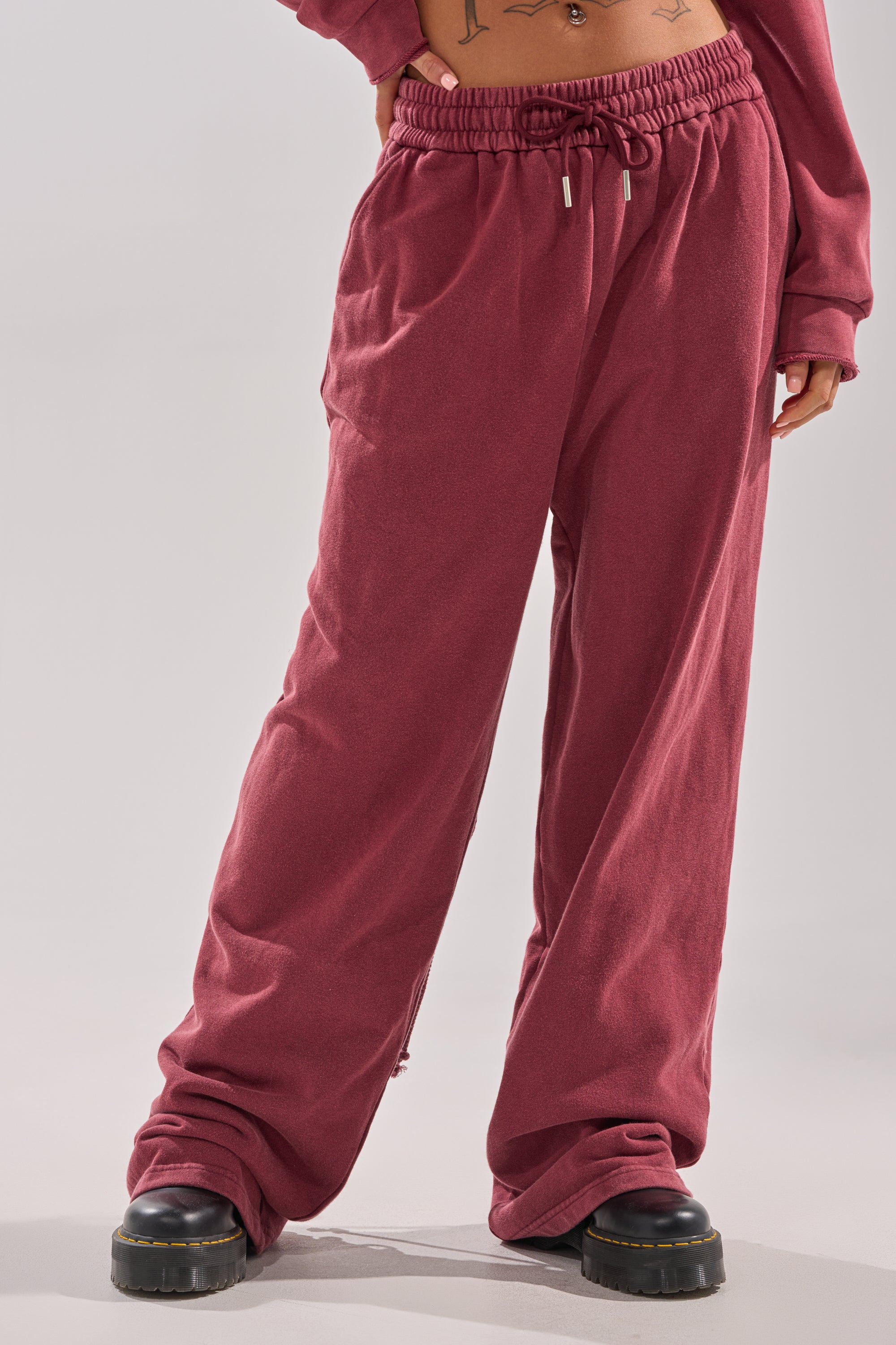 Wearing the LACED UP FOR GOOD JOGGER IN PLUM, a person with black platform shoes stands against a light background; their sweatshirt sleeves cover their hands, revealing part of a tattooed midriff.