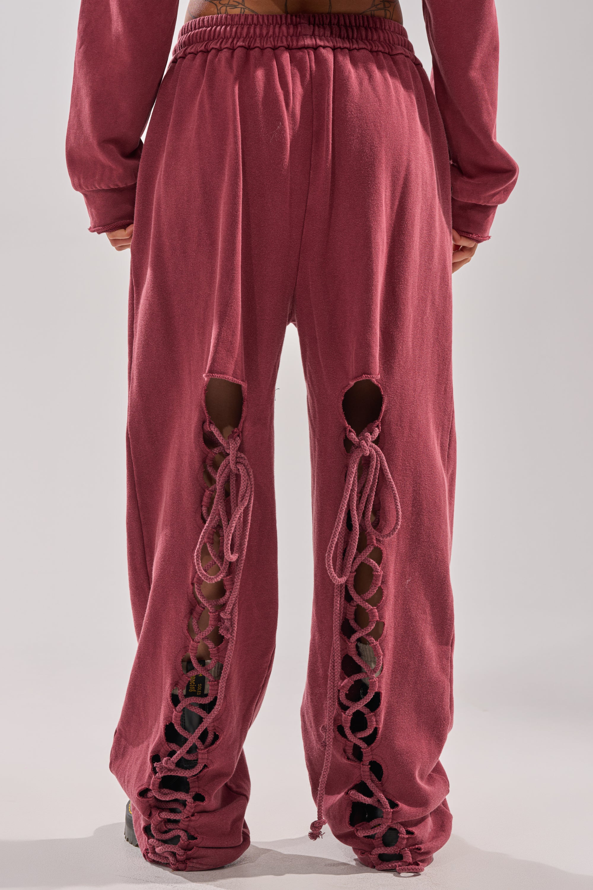 A person stands facing away, wearing the LACED UP FOR GOOD JOGGER IN PLUM with open crisscross cutout details on the back of both legs, paired with matching long sleeves.