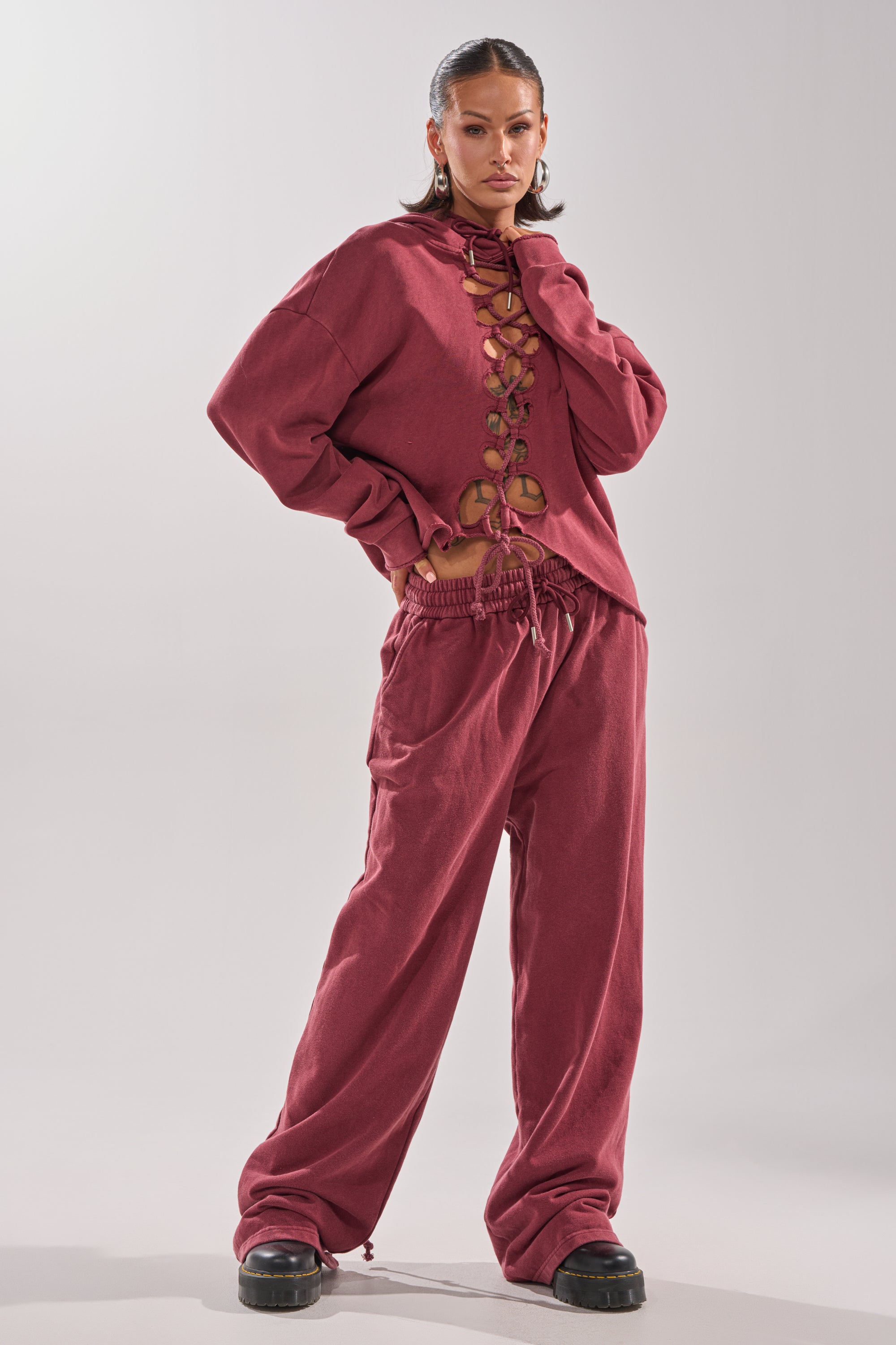 A woman stands confidently in the LACED UP FOR GOOD JOGGER IN PLUM, a dark rose lace-up set with a stylish front-tie top and matching wide-leg pants. She completes her look with hoop earrings, sleek hair, and black platform shoes against a plain background.