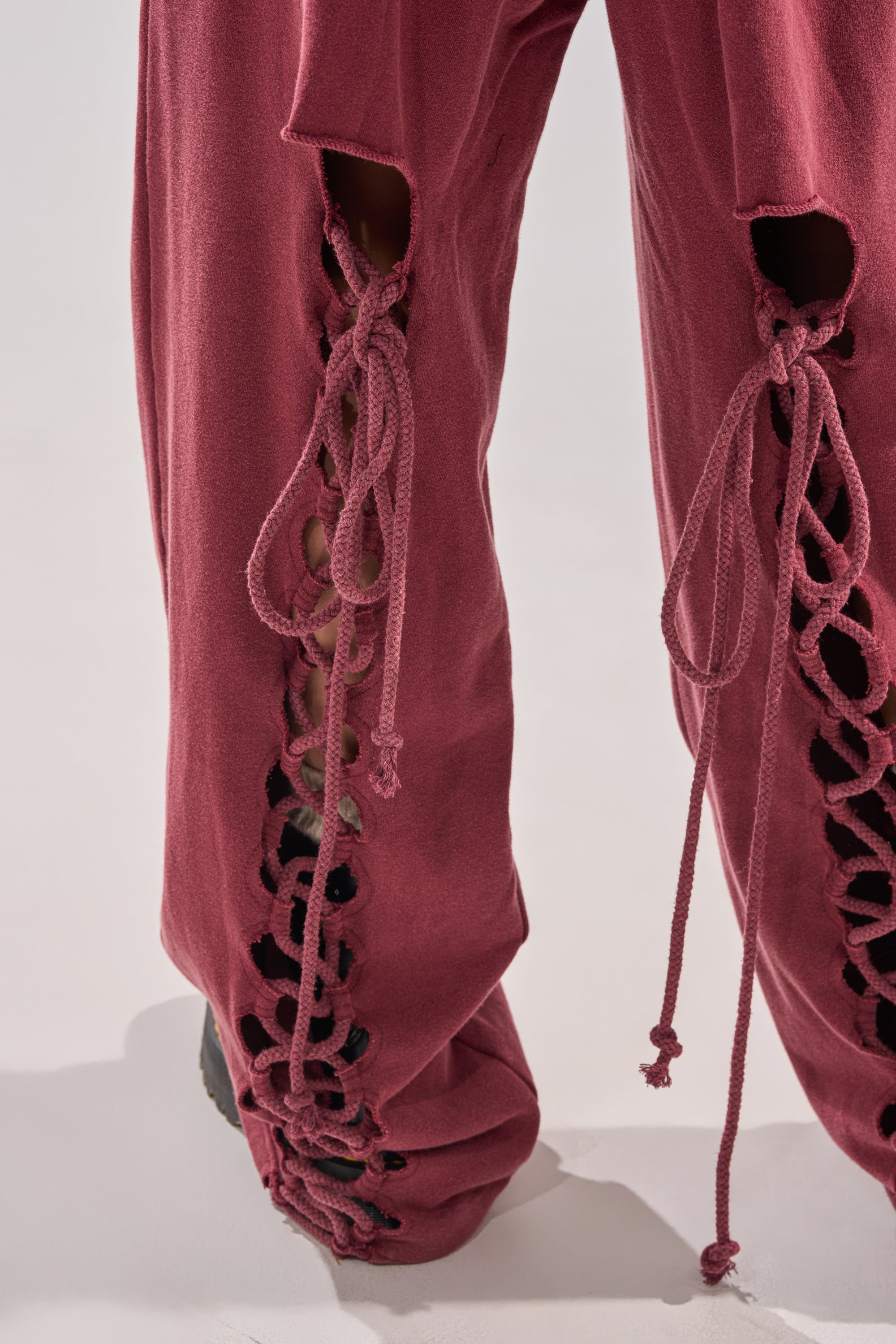 The LACED UP FOR GOOD JOGGER IN PLUM is a high-rise, loose-fitting jogger featuring front cut-out details with crisscross drawstring laces that reveal skin, extending over black shoes.