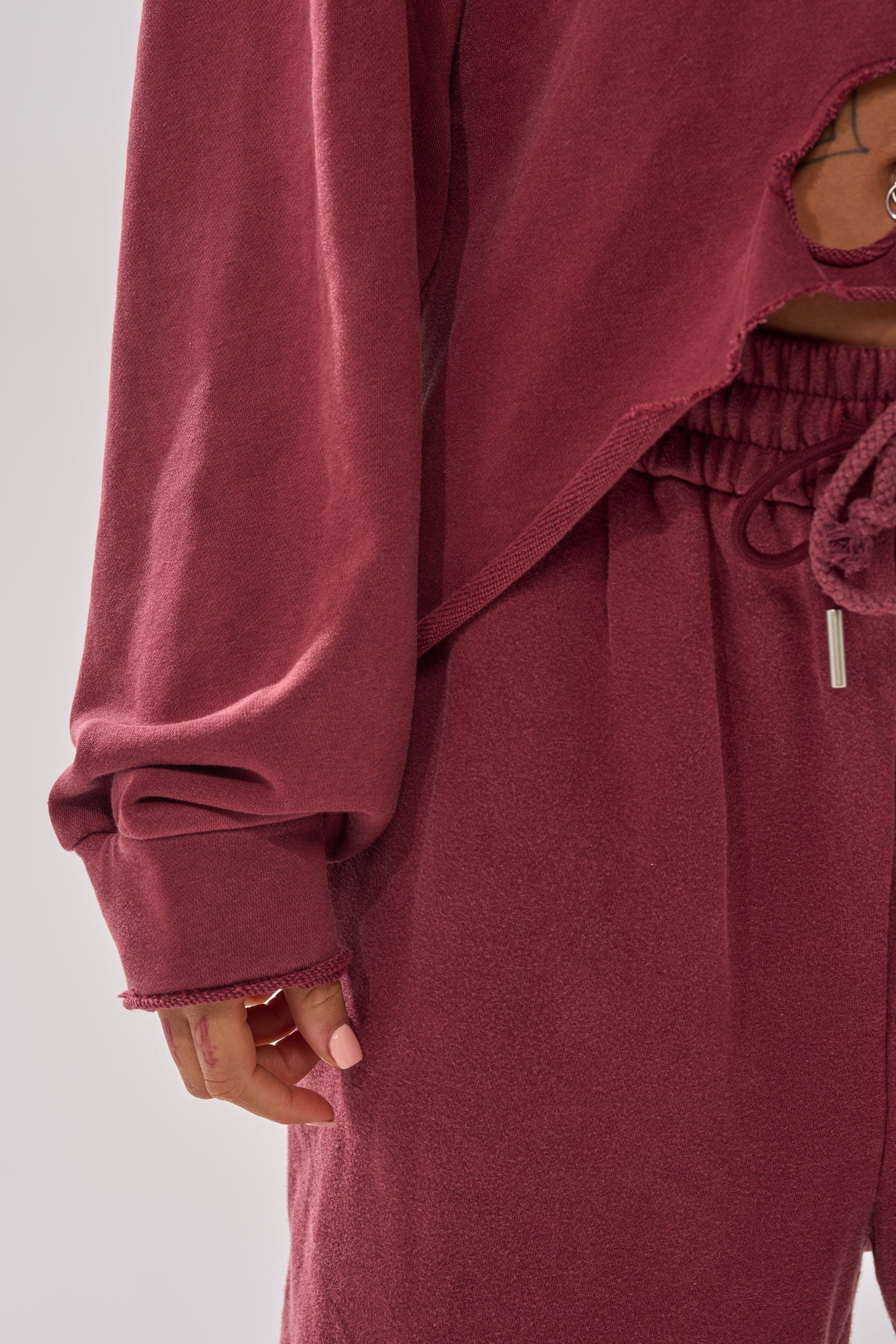 A person wears the LACED UP FOR GOOD SWEATSHIRT IN PLUM with matching drawstring pants, showing their hand with light pink nail polish. The relaxed fit offers ultimate comfort, perfect for lounging.