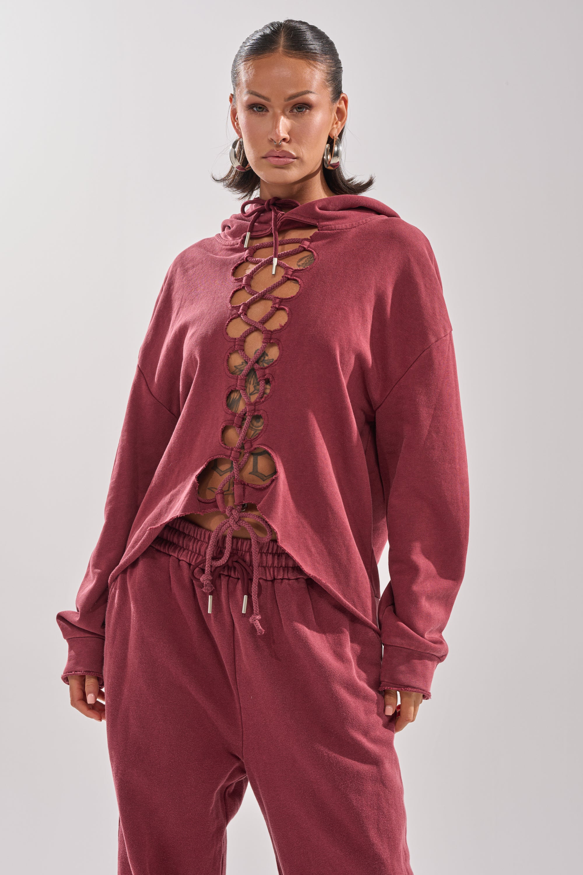 A woman confidently wears the LACED UP FOR GOOD SWEATSHIRT IN PLUM, an oversized loungewear top with a bold crisscross lace-up front that reveals her midriff as she stands against a plain background.