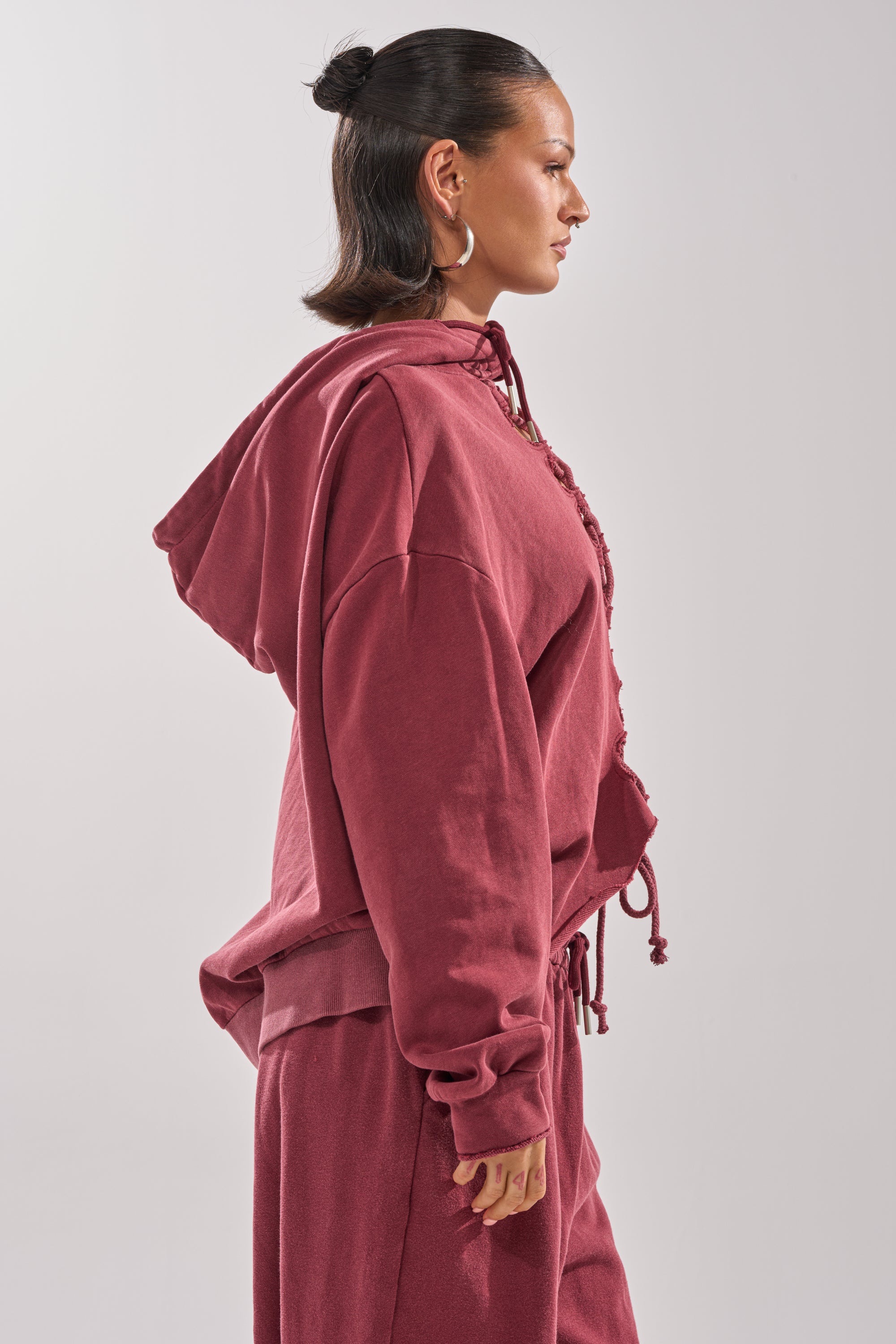 A woman stands in profile, hair pulled back, wearing the LACED UP FOR GOOD SWEATSHIRT IN PLUM with matching pants. Gold hoop earrings and a plain light background highlight her relaxed, stylish look.