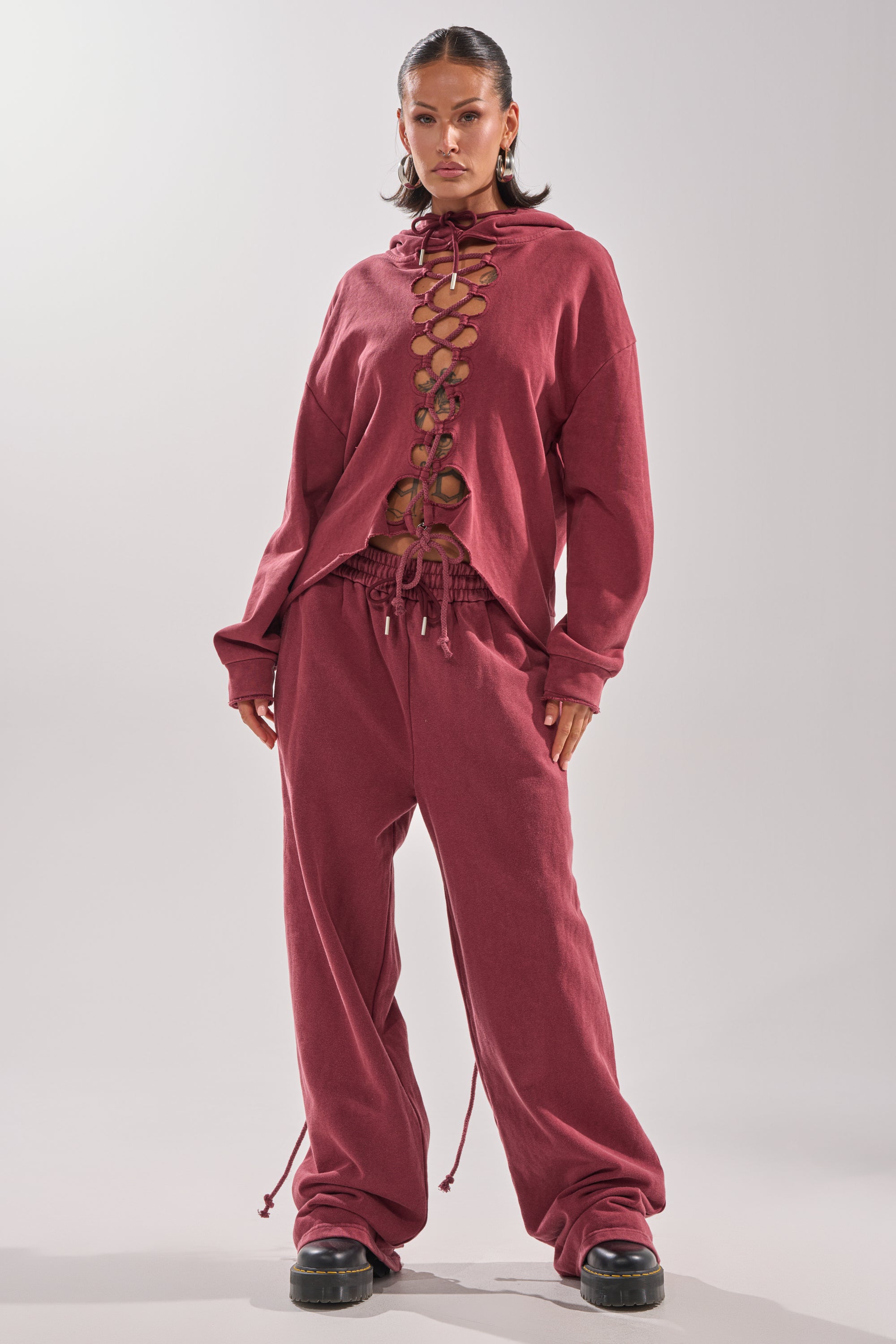 A woman stands confidently in the LACED UP FOR GOOD SWEATSHIRT IN PLUM—an oversized sweatshirt with front cutouts—paired with matching baggy pants and black platform shoes against a plain light background.