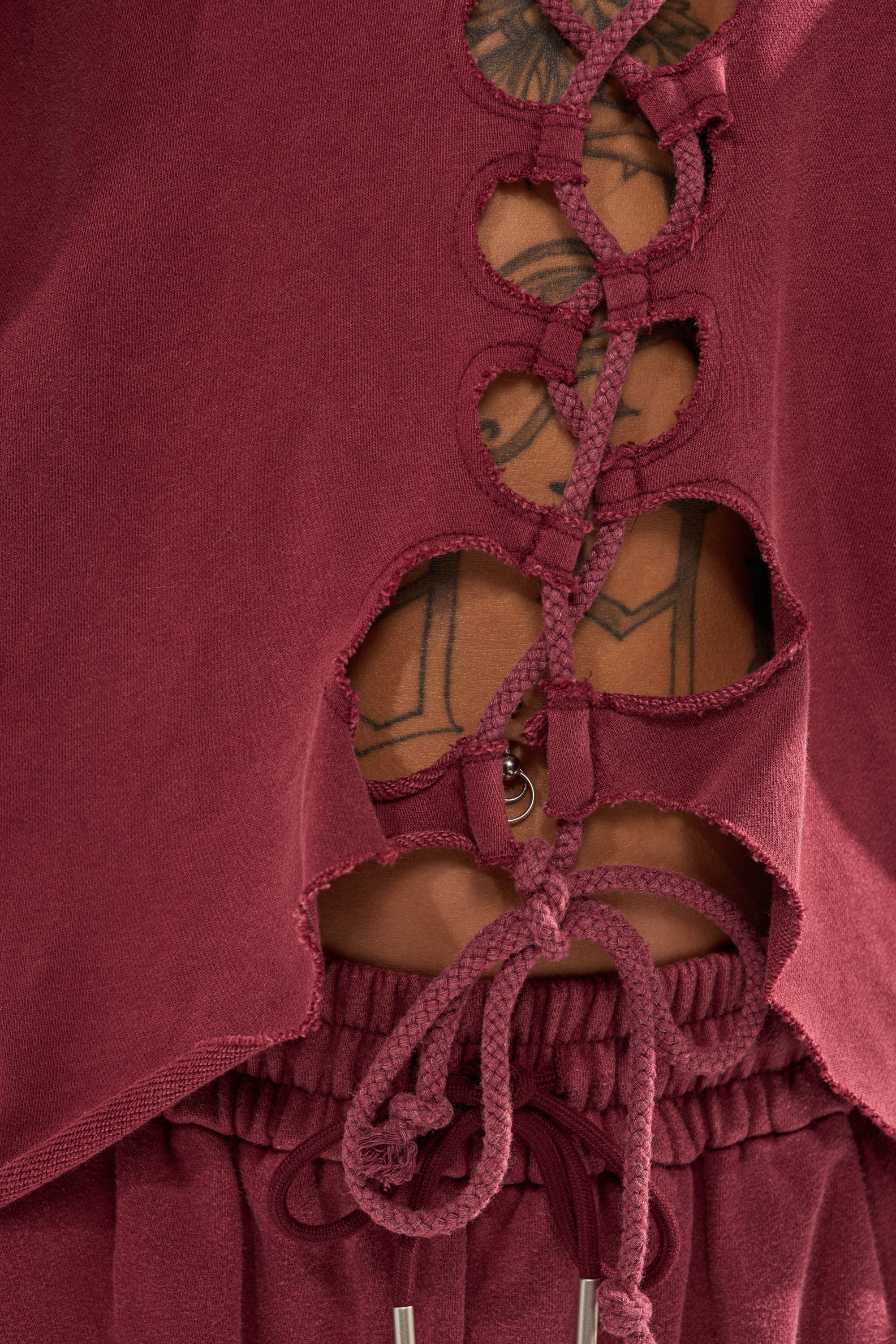 A close-up of the LACED UP FOR GOOD SWEATSHIRT IN PLUM shows lace-up detailing, drawstrings, and unfinished edges, revealing part of a tattooed torso with geometric shapes.