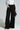 A person wearing the FREYA ZIPPER PANT, paired with a shiny silver jacket, holds a small reflective clutch. With one hand in their pocket and open-toed shoes adorning their feet, they stand against a neutral background.