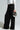 A person in a silver blouse and the black FREYA ZIPPER PANT, which is a high-rise wide-leg trouser, holds a silver crescent-shaped handbag. The outfit is stylish and modern, highlighting the contrast between the shiny top and the dark pants with zipper embellishments.