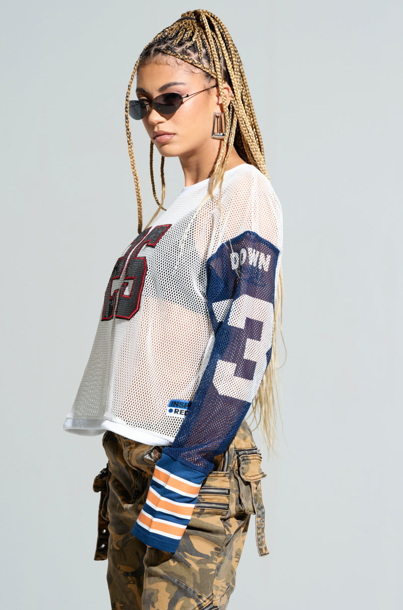 A person with long braided hair wears sunglasses and the WINNERS EVERYWHERE TOP, an oversized mesh jersey adorned with sequined "25" and "Brown" details, paired with camouflage pants. They stand in a confident pose against a plain background.