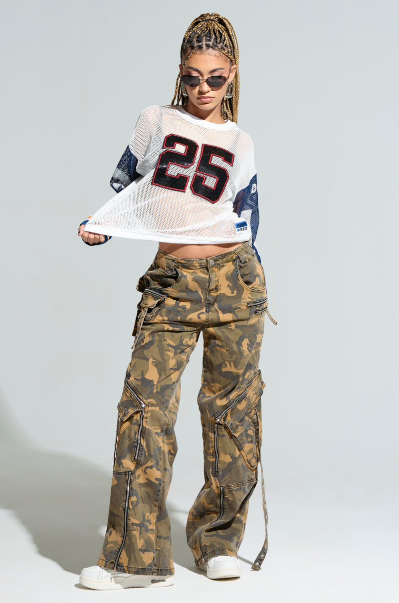 A person with braided hair, sunglasses, and hoop earrings confidently stands against a plain background, lifting their oversized white mesh jersey top emblazoned with the number 25. They pair the "WINNERS EVERYWHERE TOP" with camo cargo pants and white sneakers.