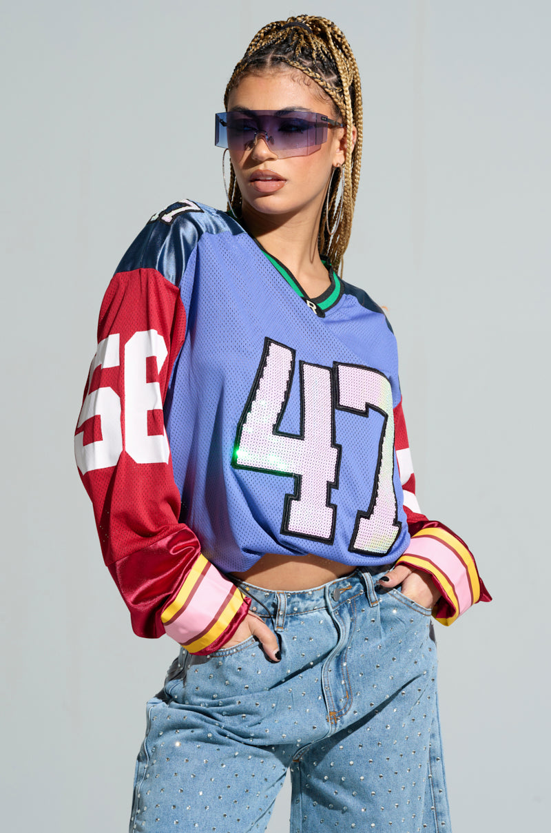 A person confidently stands in a vibrant color block pattern, showcasing a blue GAME TIME JERSEY featuring the number 47 and red and blue sleeves. Their braided hair and large sunglasses complement the oversized silhouette, which is paired with ripped denim jeans. 0