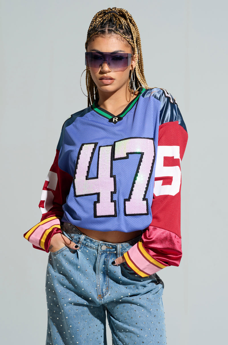 A woman confidently stands with her hands in her jean pockets, wearing oversized sunglasses and the GAME TIME JERSEY, featuring the number 47. Her braided hair complements the jersey's vibrant maroon and pink color block design. The plain background accentuates her bold style. 1